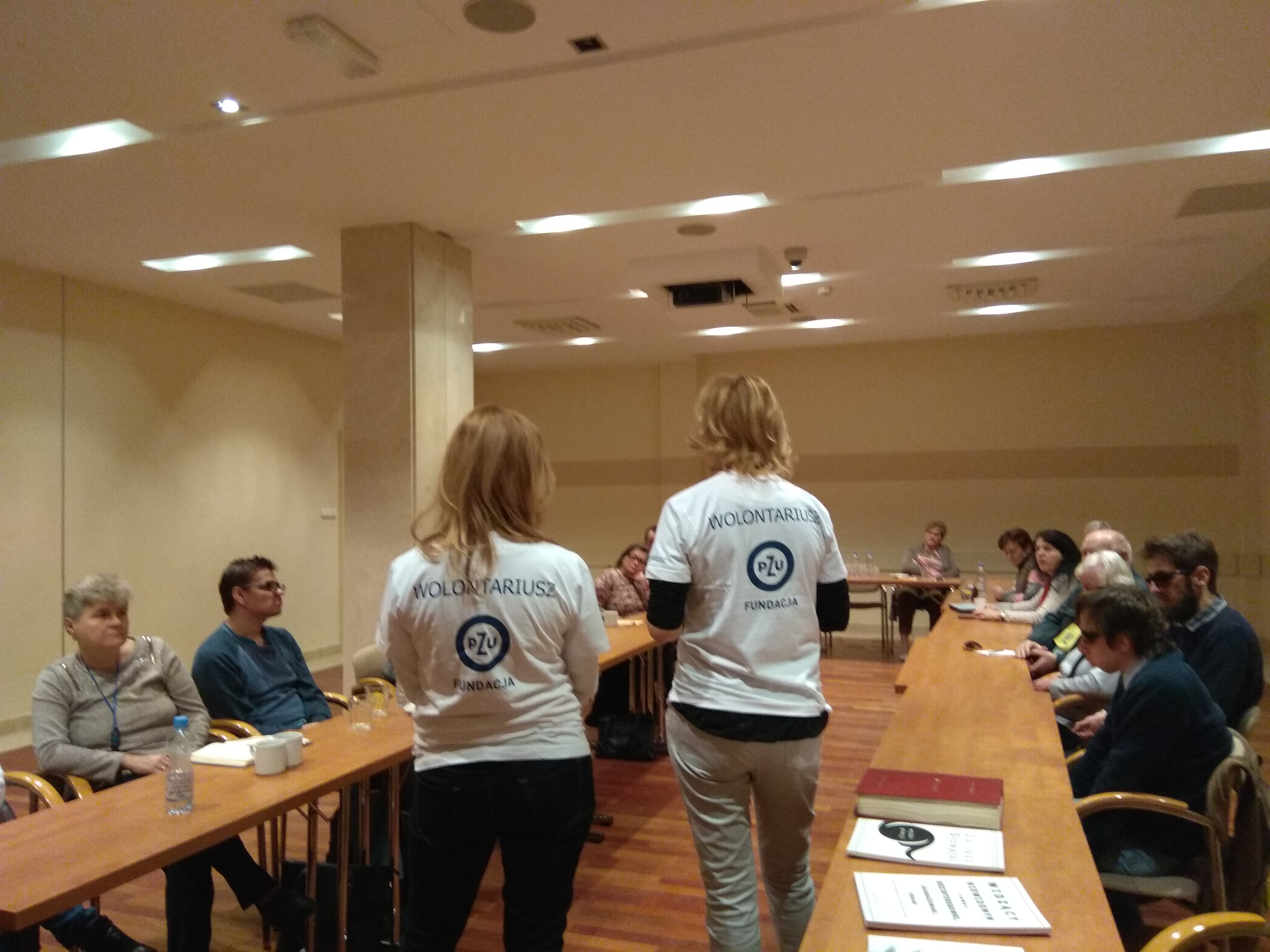 W jasnej przestrzeni sali konferencyjnej widoczni uczestnicy zjazdu siedzący po bokach sali, a w środku między stołami stojące dwie panie, obie blondynki,  w białych koszulkach widziane od tyłu, na koszulkach logo Fundacji PZU, jej nazwa i napis Wolontariusz.